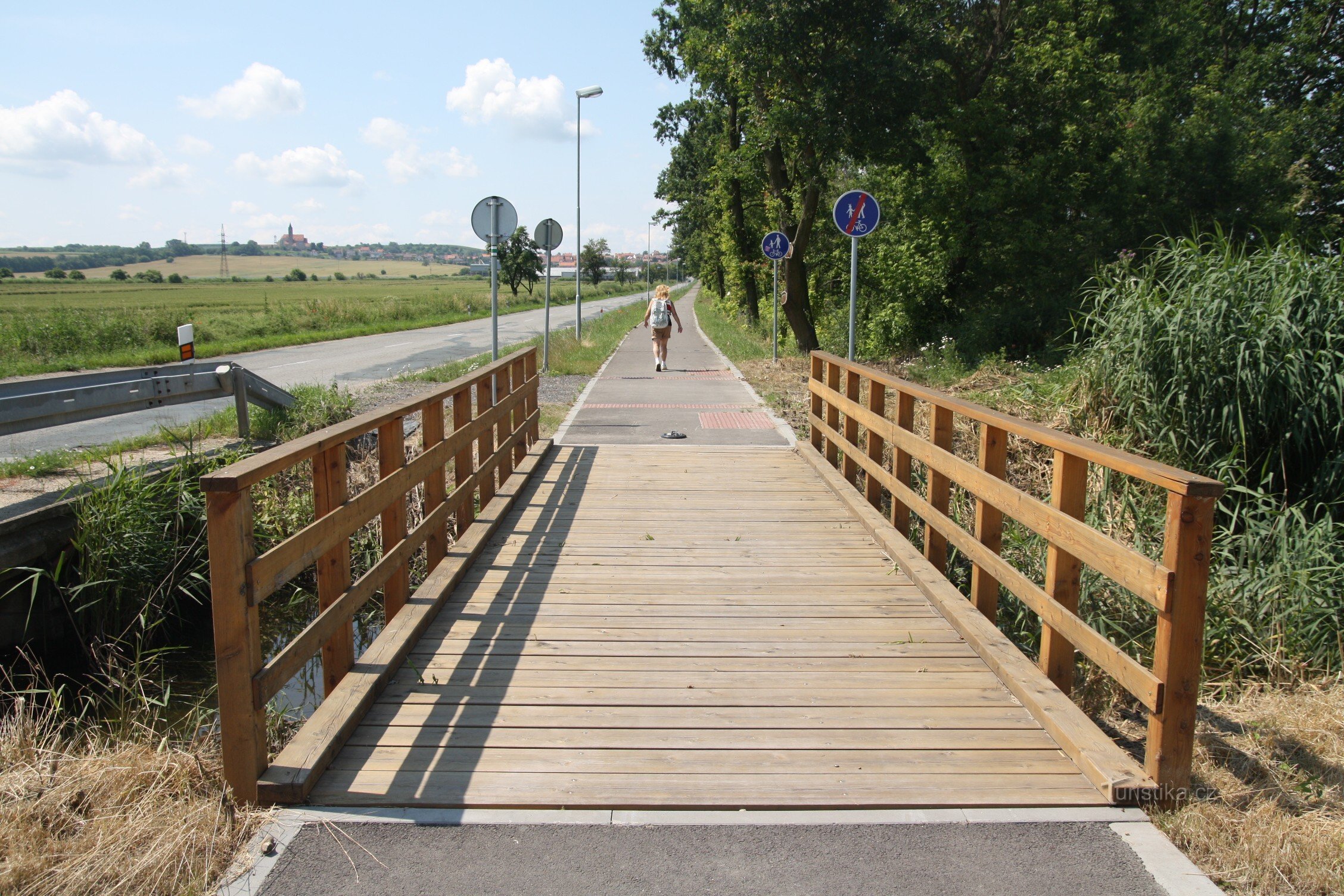 Am Ende des Weges eine Brücke über den Bach, im Hintergrund am Horizont die dominante Jaječ-Kirche