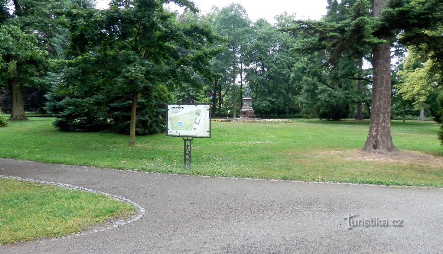 Placa de orientação com a designação de árvores e arbustos plantados, ao fundo a escultura de Mateřsk