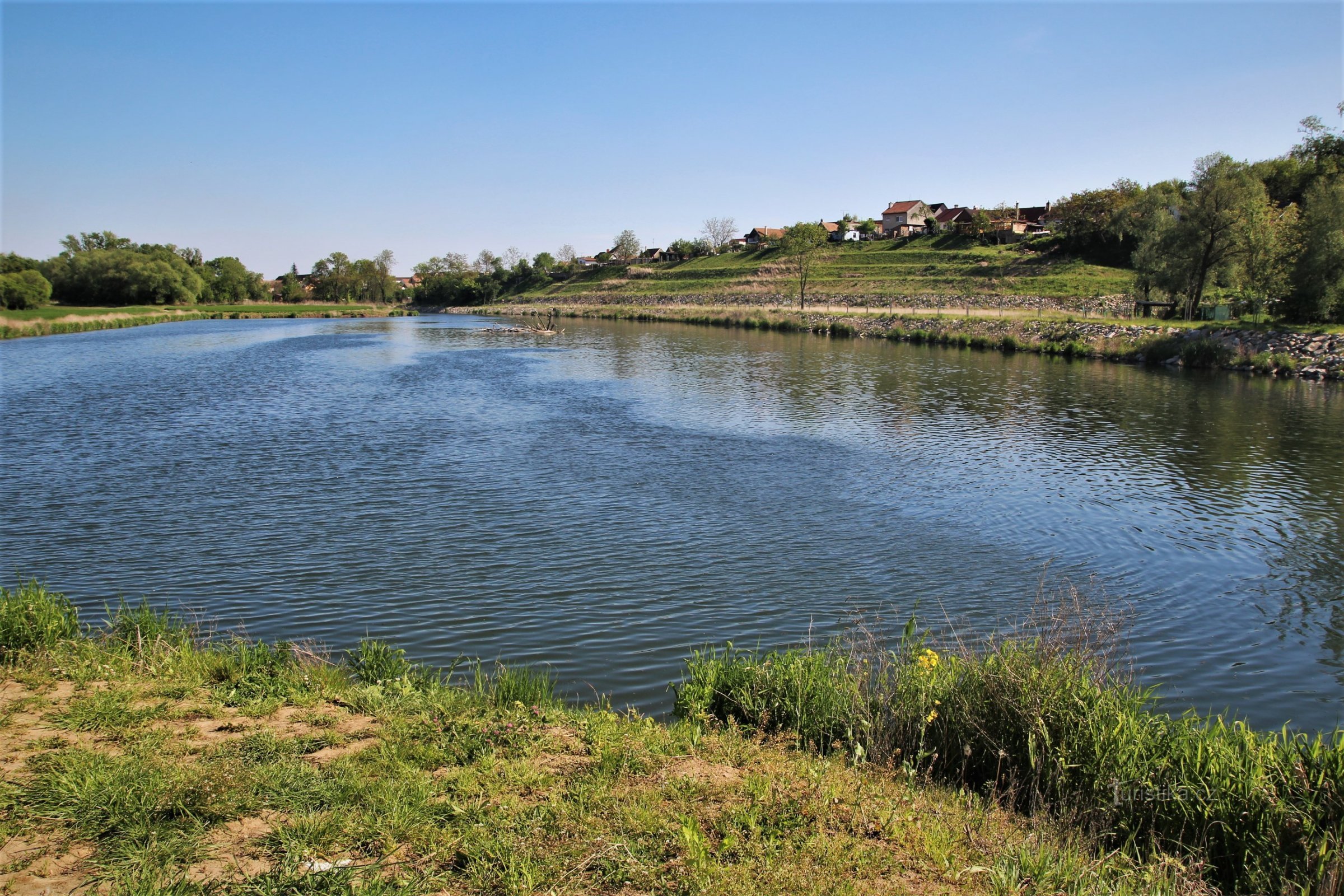 Uma vasta piscina de água e acima dela terraços de uma área séria, ao fundo a vila de Bulhary