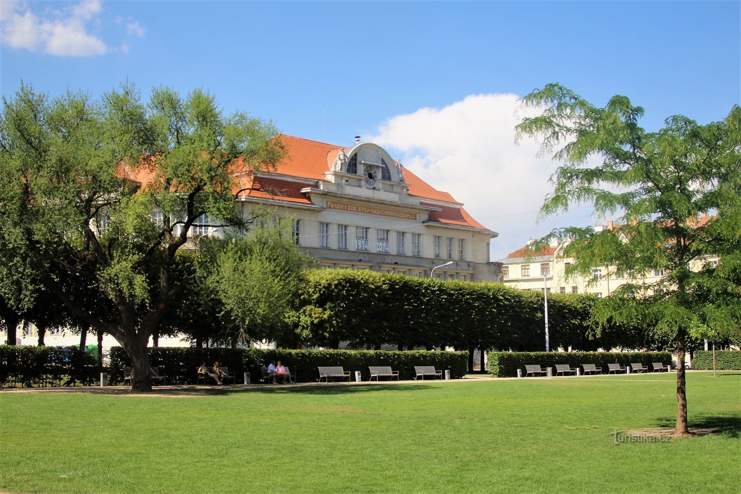 In the foreground is a grassy area of ​​the park framed by a row of trees, in the background the building of the Elementary School